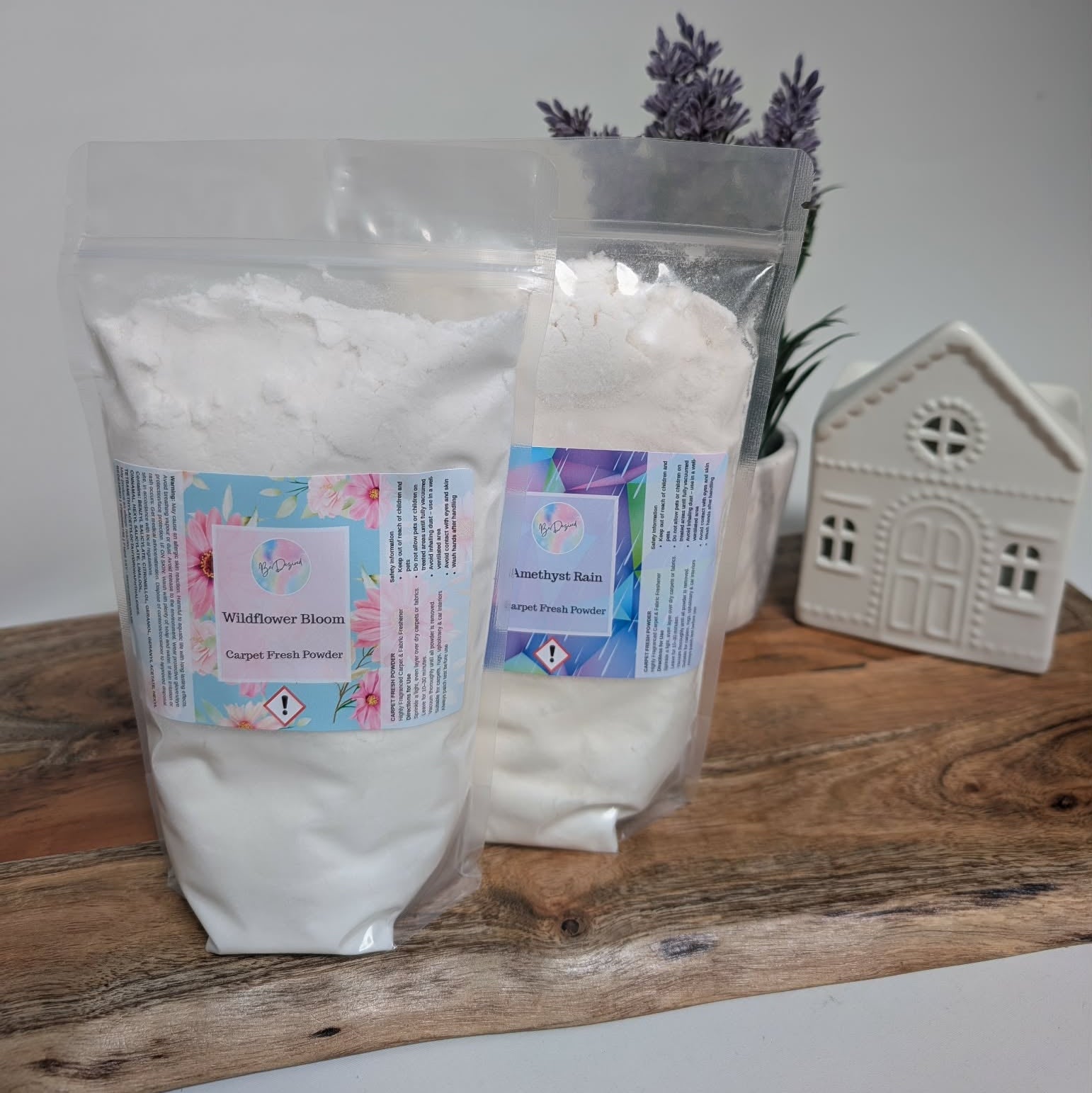 Two packages of white carpet fresh powder with labels on a wooden surface, lavender flowers, and a small house decoration in the background.
