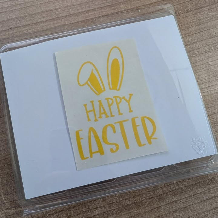 Happy Easter card with yellow text and bunny ears design on a white card, placed on a wooden surface. For Home Bargain Style Boxes